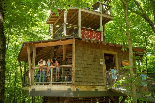A wooden treehouse surrounded by lush green trees, with people enjoying the view from the balcony.