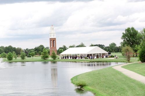 A large white tent by a pond, with a clock tower in the background and trees lining the area under a cloudy sky.