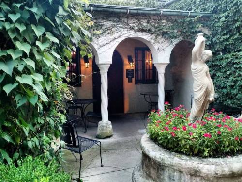 A serene courtyard with ivy-covered walls, a statue, and blooming flowers, featuring arched columns and outdoor seating.