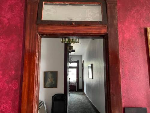 A doorway with a wooden frame leads to a hallway with red walls and a carpet, featuring a chandelier and artwork.