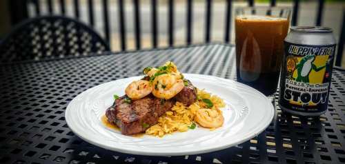 A plate of steak and shrimp over rice, with a stout beer and a can on a black outdoor table.