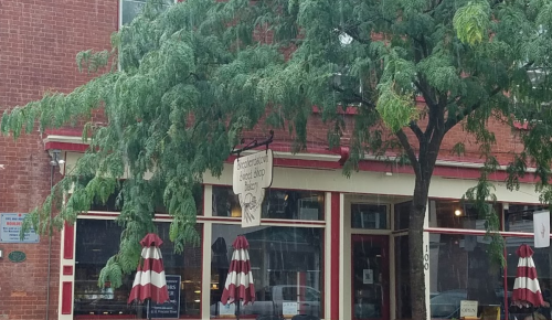 A bakery storefront with a sign, surrounded by green trees and red-and-white striped umbrellas.