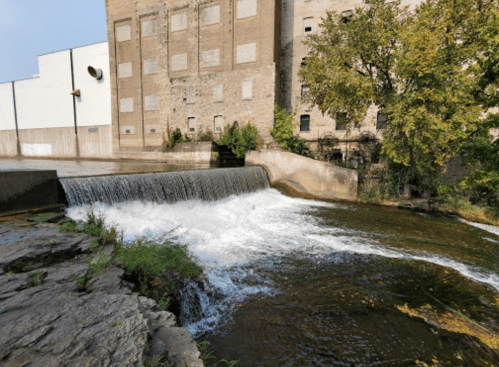 A small waterfall flows over a stone ledge beside an old industrial building, surrounded by greenery.