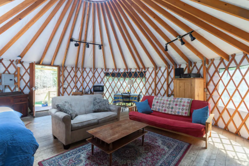 Cozy yurt interior with wooden beams, a red and beige sofa, a coffee table, and a dining area. Natural light from windows.