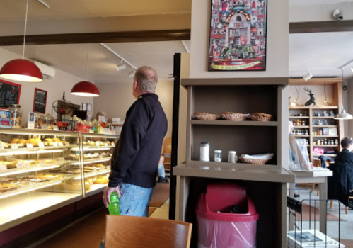 A man stands in a café, looking at a display case filled with pastries, with seating and decor in the background.
