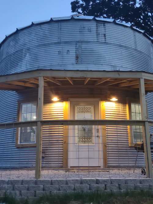 A round, metal-clad house with a porch, illuminated by two lights, set against a twilight sky.