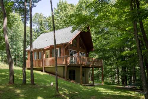 A cozy wooden cabin nestled among trees, featuring a deck and large windows, set in a lush green landscape.