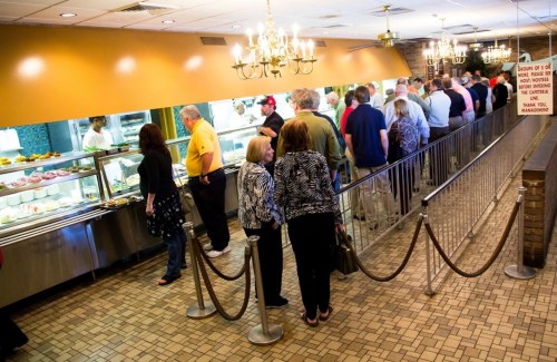 A long line of people waiting at a buffet, with a dessert display and chandeliers in a restaurant setting.