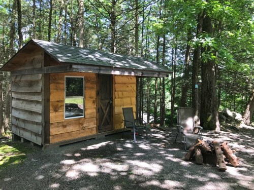 A rustic wooden cabin surrounded by trees, with two chairs and a stack of firewood in front.