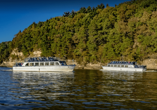 Two white boats glide through calm waters, surrounded by lush green trees and rocky cliffs.