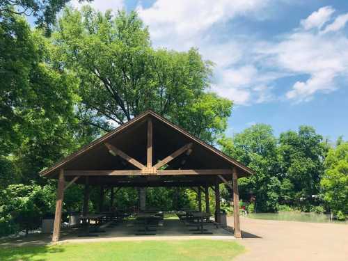 A wooden pavilion with picnic tables, surrounded by lush green trees and a blue sky with scattered clouds.