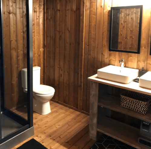 A rustic bathroom featuring wooden walls, a shower, a toilet, and a double sink with a wooden vanity.
