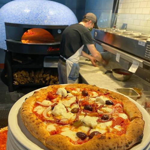 A freshly baked pizza topped with cheese and olives, with a chef preparing dough in the background near a wood-fired oven.