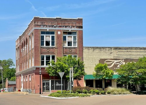 Historic brick building labeled "Masonic Temple" next to a storefront, with trees and a clear blue sky in the background.