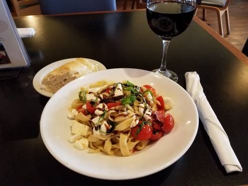 A plate of pasta with cherry tomatoes and cheese, accompanied by a glass of red wine and a bread roll on the side.