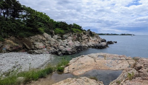 Rocky coastline with greenery, calm water, and a cloudy sky in the background. A sailboat is visible in the distance.