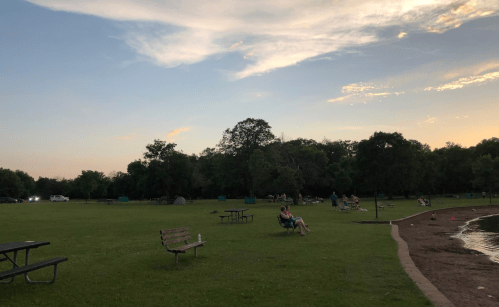 A peaceful park scene at sunset, featuring green grass, benches, and trees, with a calm water edge in the background.