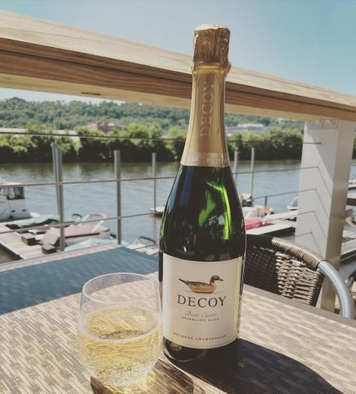 A bottle of Decoy sparkling wine next to a glass filled with wine, overlooking a scenic waterfront view.