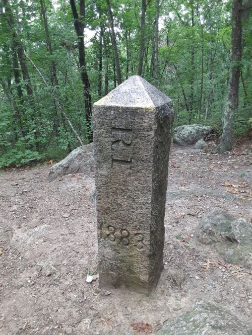 A stone marker with "RI" and "1883" engraved, surrounded by trees and rocky terrain.