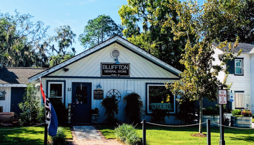 A charming white building with a sign reading "Bluffton General Store," surrounded by greenery and a welcoming entrance.