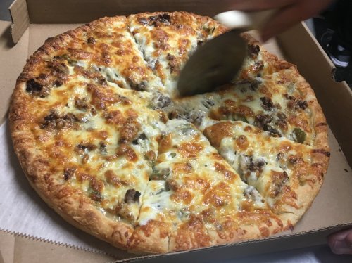 A freshly baked pizza with melted cheese and toppings, being sliced with a pizza cutter in a cardboard box.