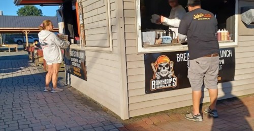 A food vendor serves a customer at a small outdoor eatery with a sign for "Bonehead's Grille."