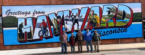 A group of five people stands in front of a colorful mural that reads "Greetings from Hayward, Wisconsin."