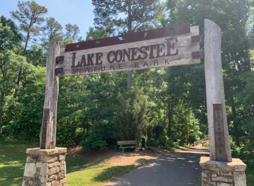 Sign at the entrance of Lake Conestee Nature Park, surrounded by trees and a pathway leading into the park.