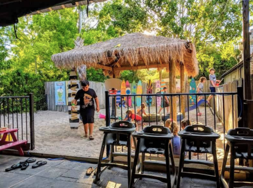 A playground with a thatched-roof structure, children playing in sand, and colorful decorations in a sunny outdoor setting.