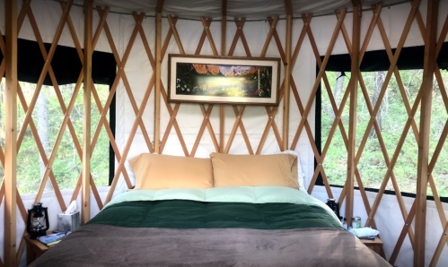 Cozy yurt interior featuring a bed with soft bedding, framed artwork, and large windows showcasing greenery outside.