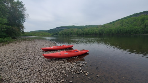 Two red kayaks sit on a rocky shore by a calm river, surrounded by lush green trees and hills.