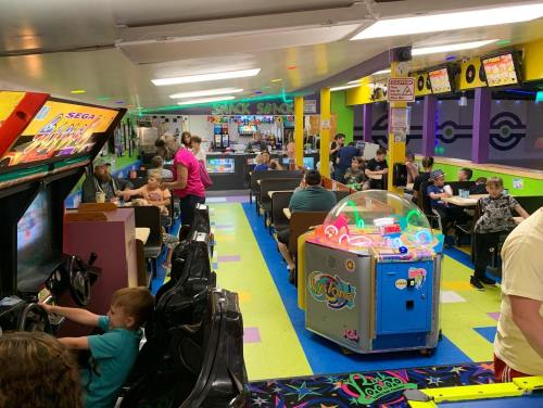 A colorful arcade filled with children and families enjoying games and snacks at tables. Bright lights and machines are visible.