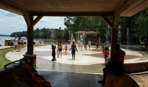 Children playing in a splash pad area with water fountains, under a shaded pavilion by a lake.