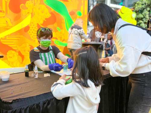 A child and an adult interact at a colorful event, with a staff member in a green apron assisting them.