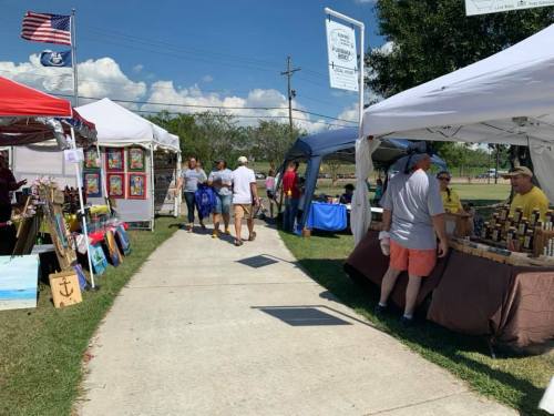 A lively outdoor market scene with vendors under tents, people browsing, and a clear blue sky above.