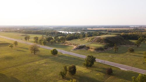 Aerial view of a grassy landscape with a hill, winding road, and a river in the distance under a clear sky.