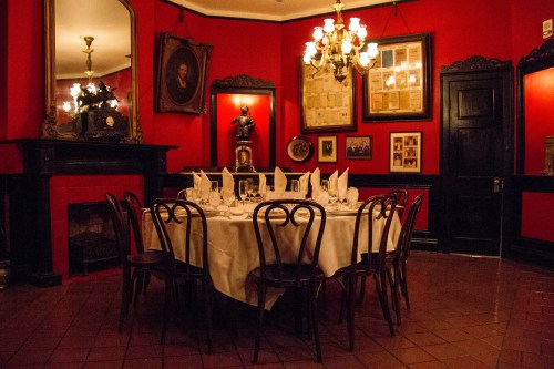 A dimly lit dining room with a round table set for dinner, surrounded by red walls and vintage decor.