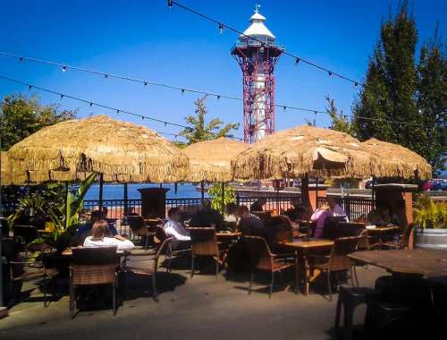 Outdoor dining area with thatched umbrellas, people seated at tables, and a lighthouse in the background under a clear blue sky.
