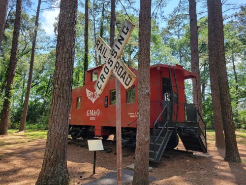 A red caboose sits among tall trees, with a railroad crossing sign in front and an informational plaque nearby.