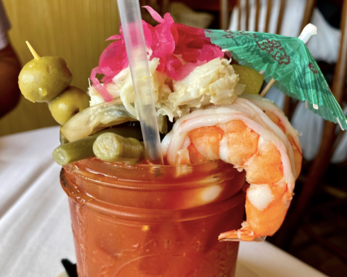 A colorful cocktail garnished with shrimp, olives, pickled vegetables, and a small umbrella in a glass.