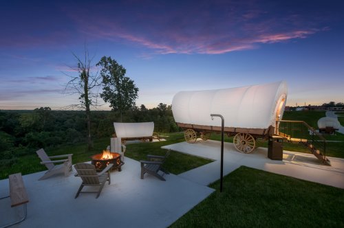 A cozy outdoor scene featuring a covered wagon, fire pit, and seating area against a twilight sky.