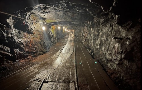 Dimly lit underground mine tunnel with wooden tracks and rocky walls, illuminated by hanging lights.
