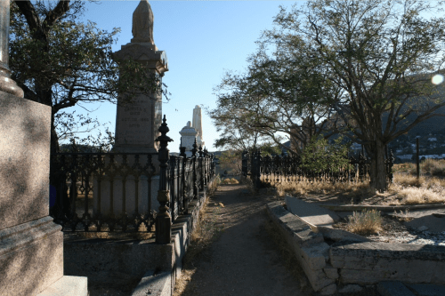 A narrow path between ornate gravestones and trees in a cemetery, with sunlight filtering through the branches.