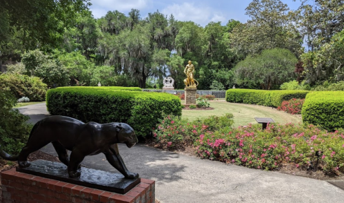 A garden scene featuring a black panther statue in the foreground and a golden statue surrounded by greenery and flowers.
