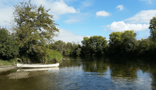 A serene river scene with a white canoe on the shore, surrounded by lush green trees and a blue sky.