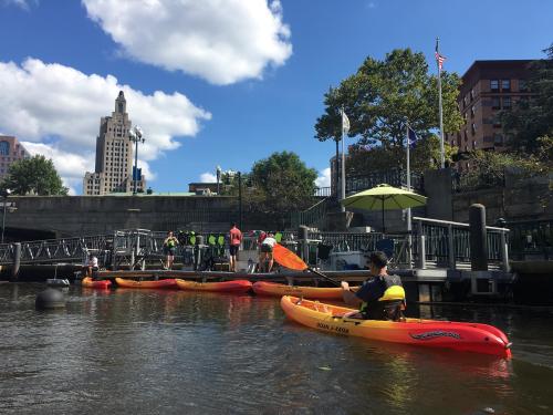 A kayaker paddles near a dock with people and buildings in the background on a sunny day.