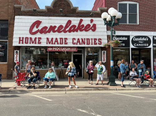 A candy shop with a vintage sign, featuring a line of people sitting outside, waiting to enter.