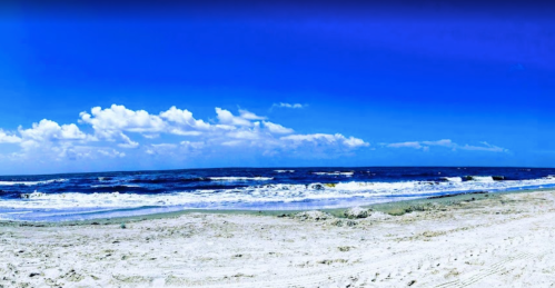 A serene beach scene with soft waves, sandy shore, and a vibrant blue sky dotted with fluffy white clouds.