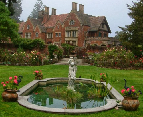 A grand mansion surrounded by lush gardens, featuring a pond with a statue and colorful flowers.