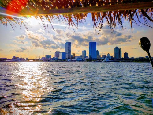 Sunset over a city skyline, viewed from the water, with reflections on the surface and clouds in the sky.